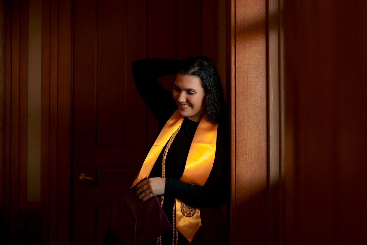 Woman in graduation regalia with gold sash posing in warm light between wooden doors.