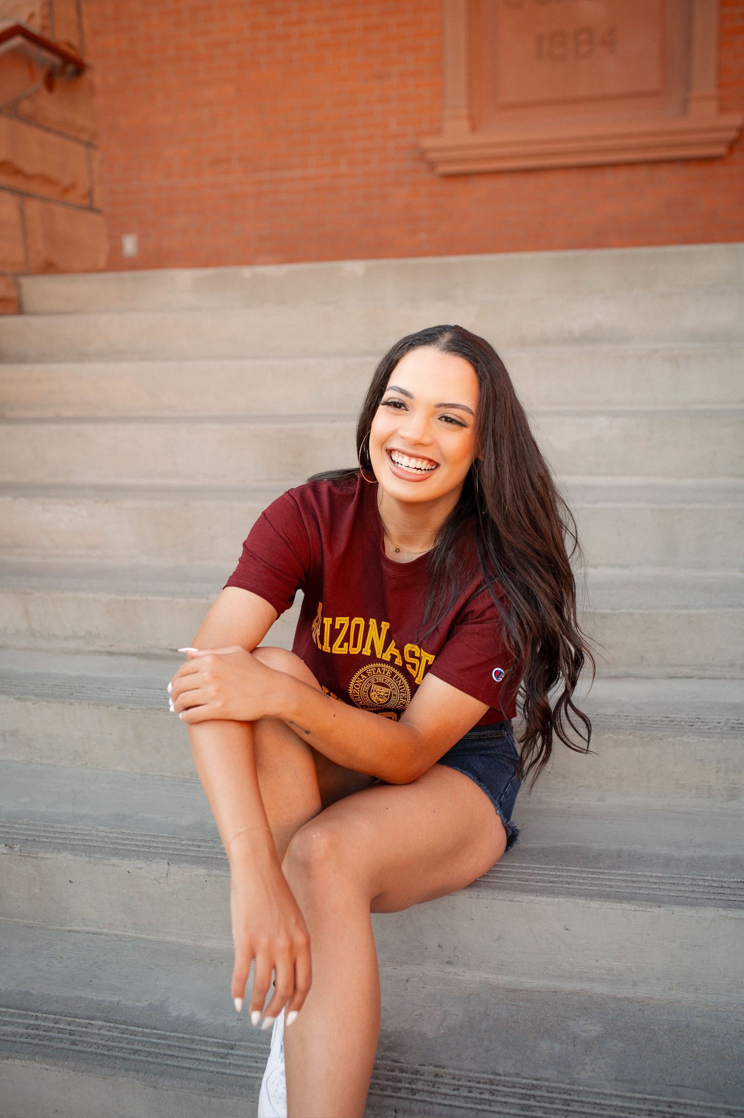 Smiling young woman in Arizona State University shirt sitting on concrete steps outside a brick building.