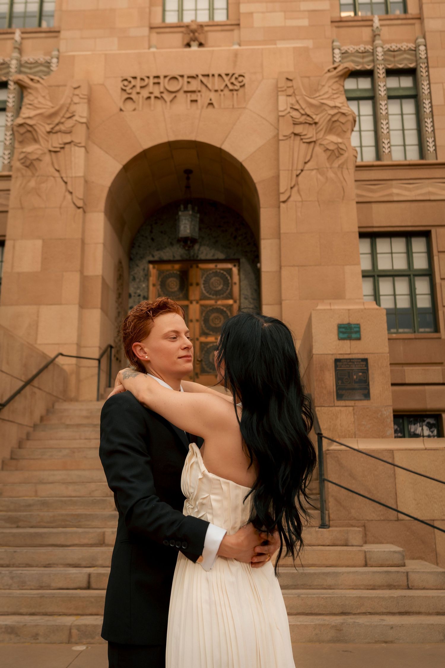 Couple embracing in wedding attire outside Phoenix City Hall, bride in white gown with long black hair, groom in black suit.