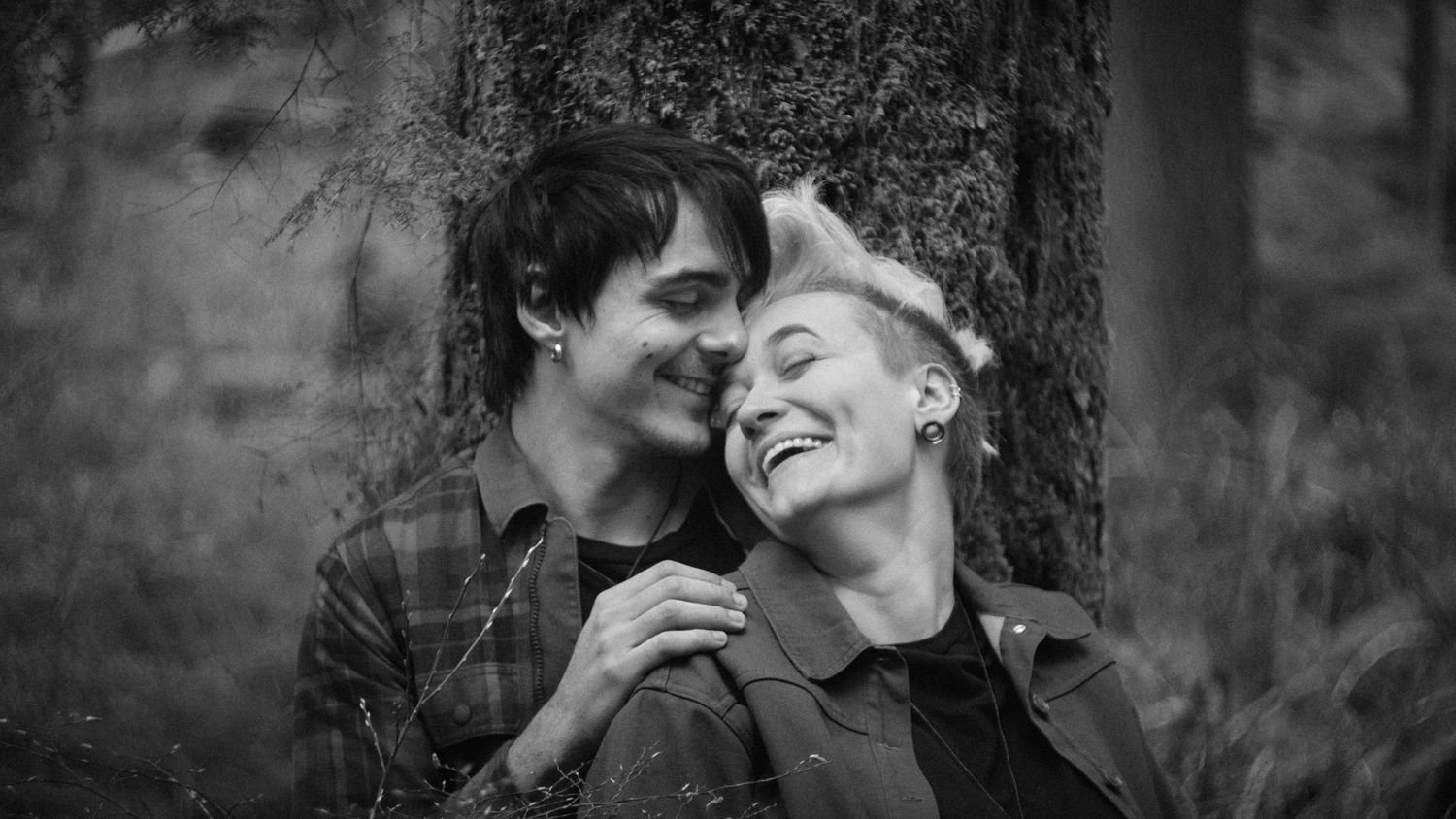 A couple laughing and embracing while leaning against a tree in a forest in Washington State. Photo by N&D Wedding Photography and Film