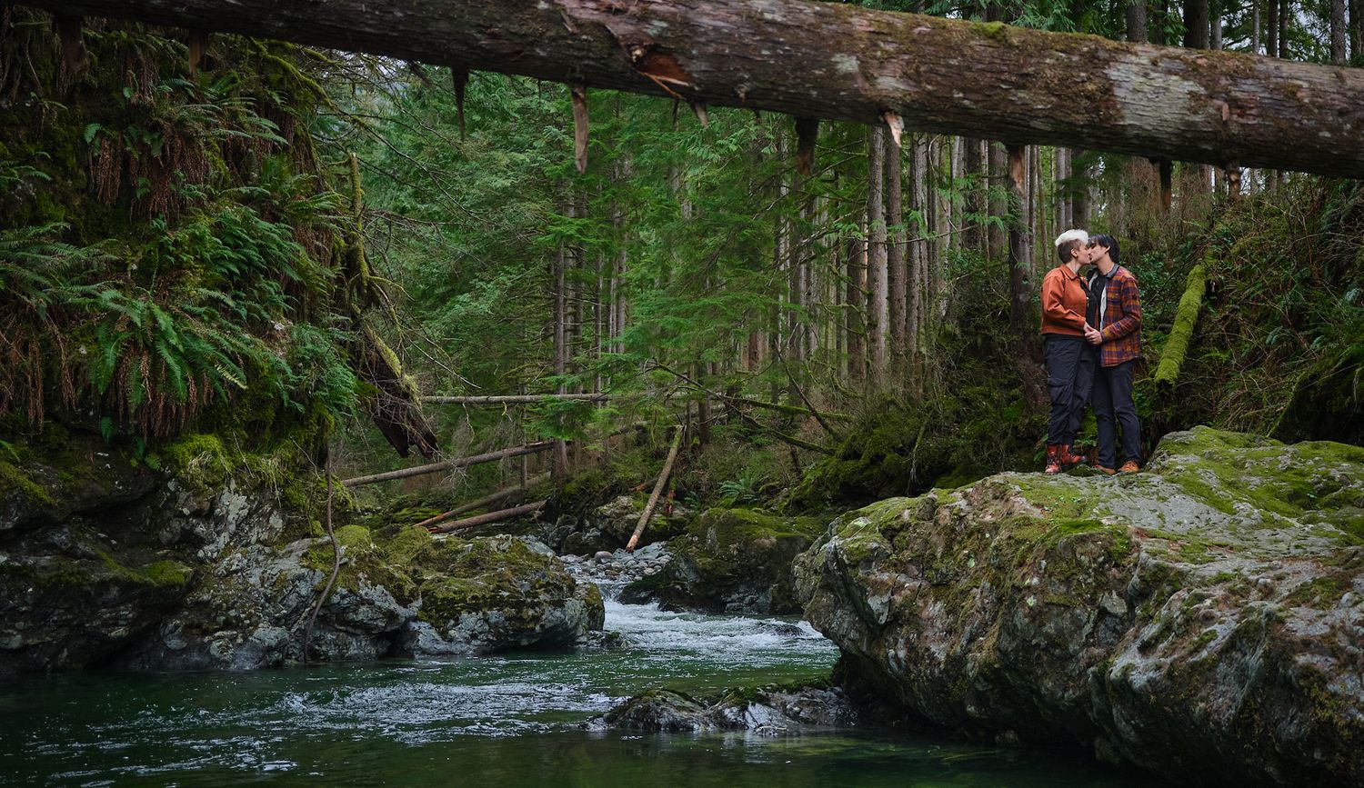A couple kissing by a river in a forest in Washington State. Photo by N&D Wedding Photography and Film