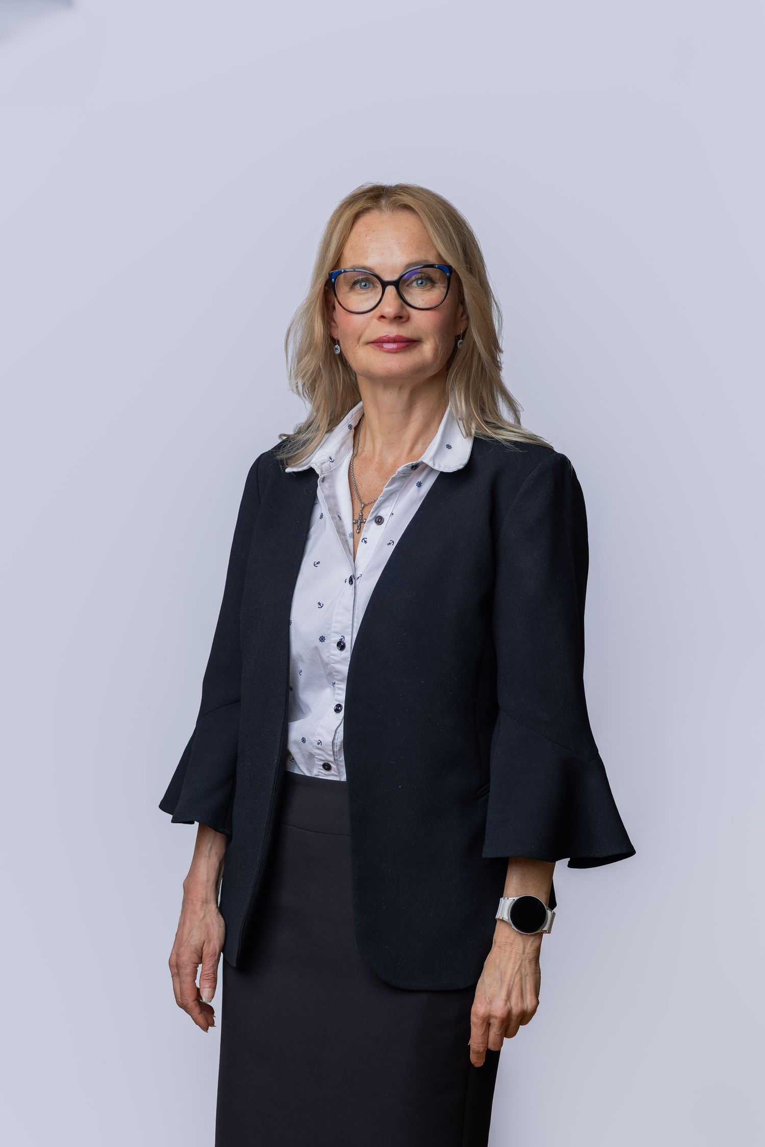 Professional woman in black blazer and glasses posing against light gray background in formal business attire.