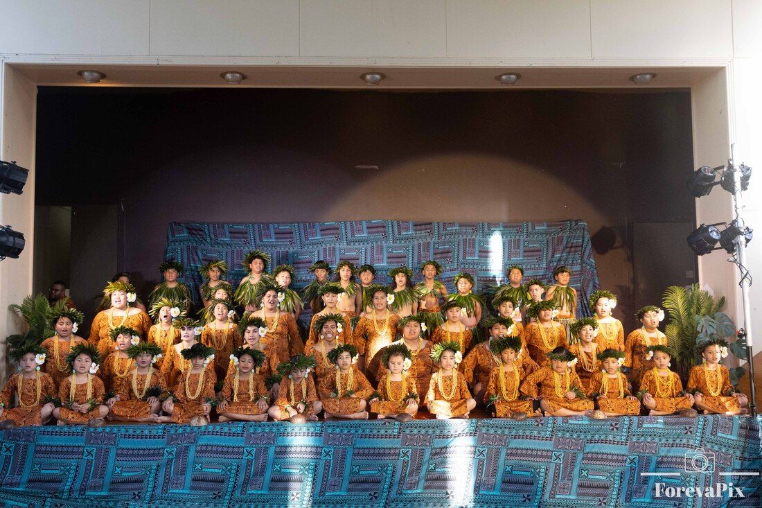 Large group of children in traditional Hawaiian costumes performing on a decorated stage at a cultural dance recital.