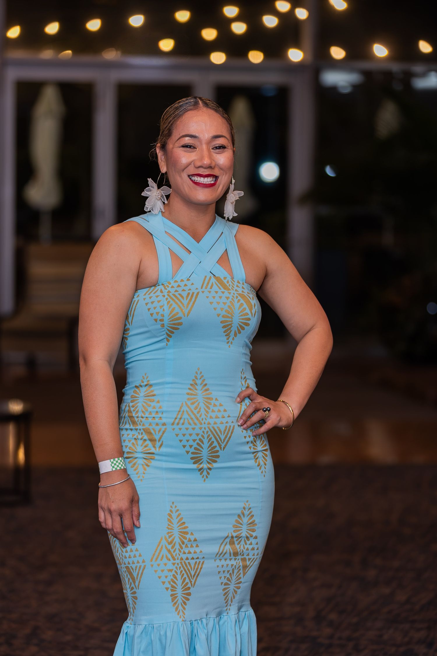 Woman in light blue and gold geometric halter dress smiling at night event with string lights.