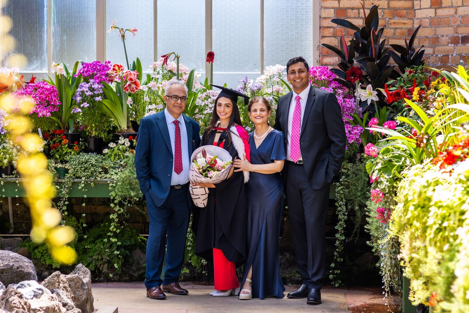Graduate in cap and gown holds bouquet, posing with three smiling guests among vibrant colorful flowers.
