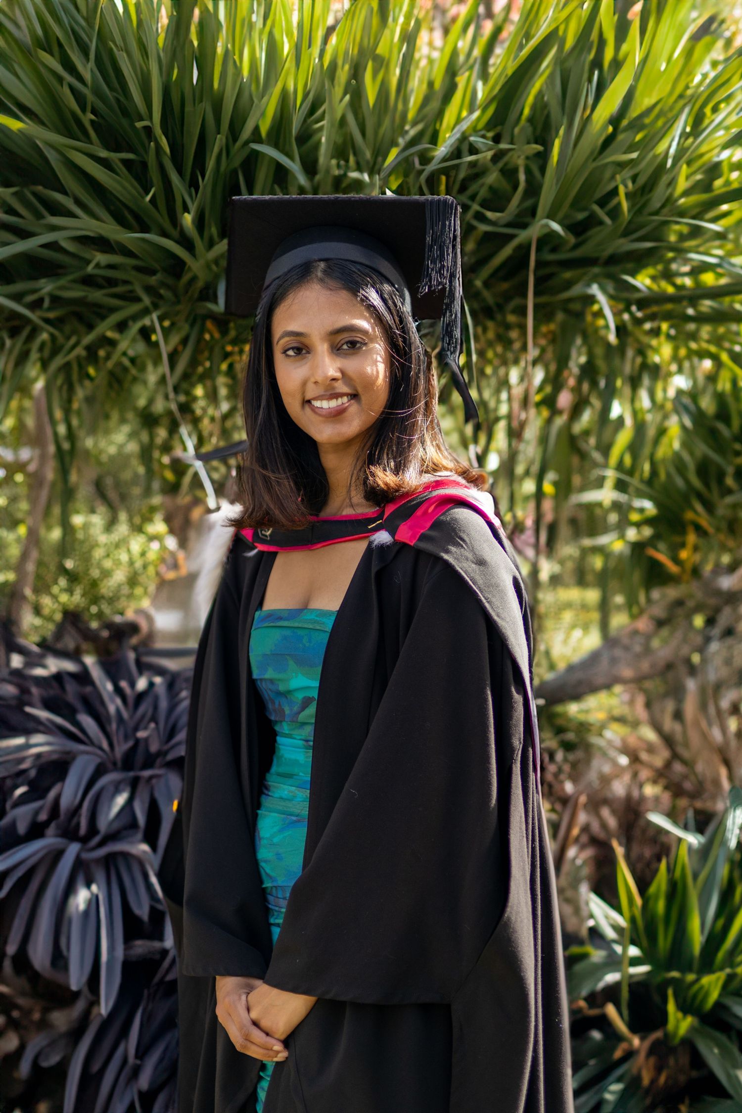 Young woman in black graduation gown and mortarboard cap smiling outdoors among tropical green plants.