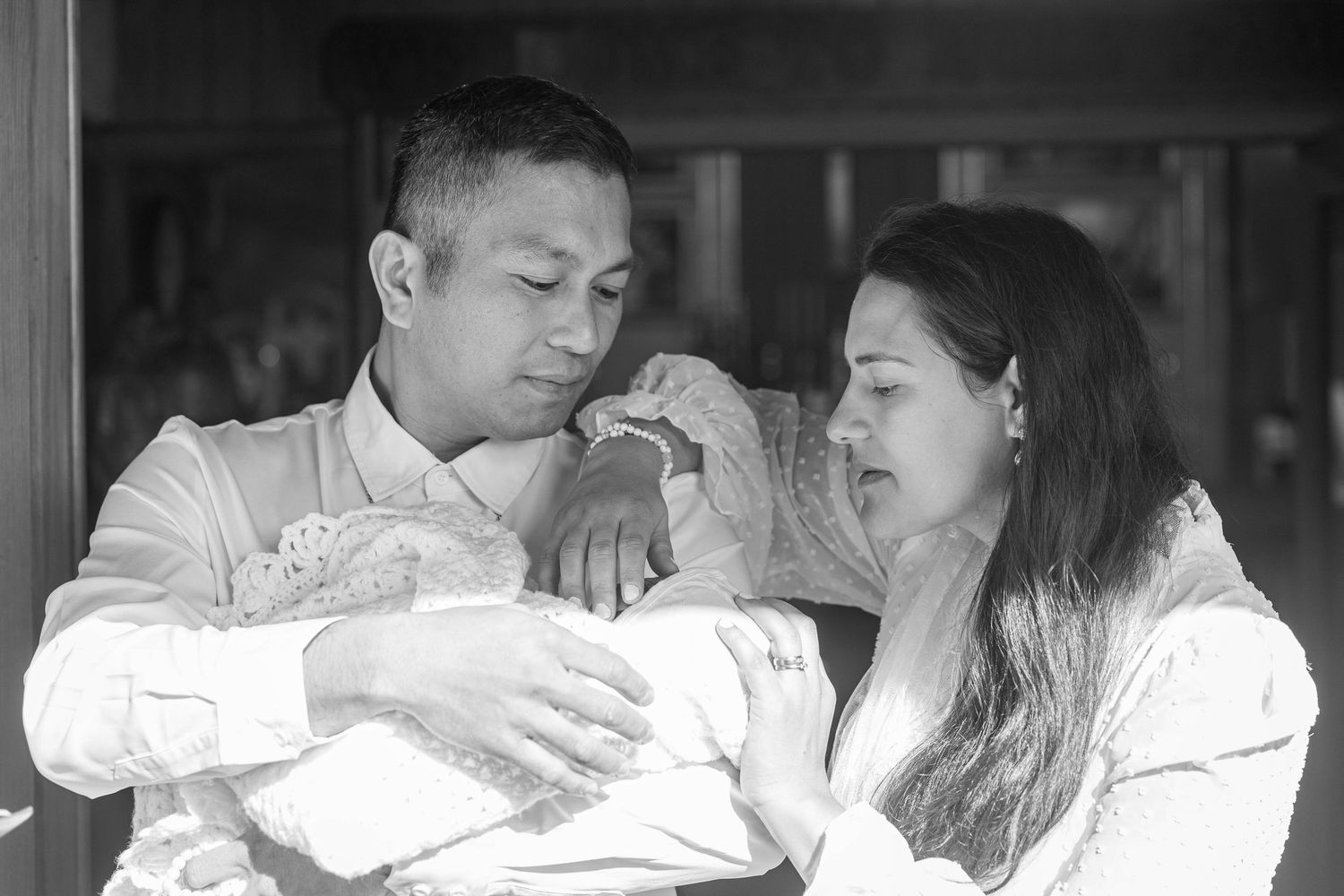 Black and white photo of new parents lovingly gazing at their newborn baby wrapped in white cloth.