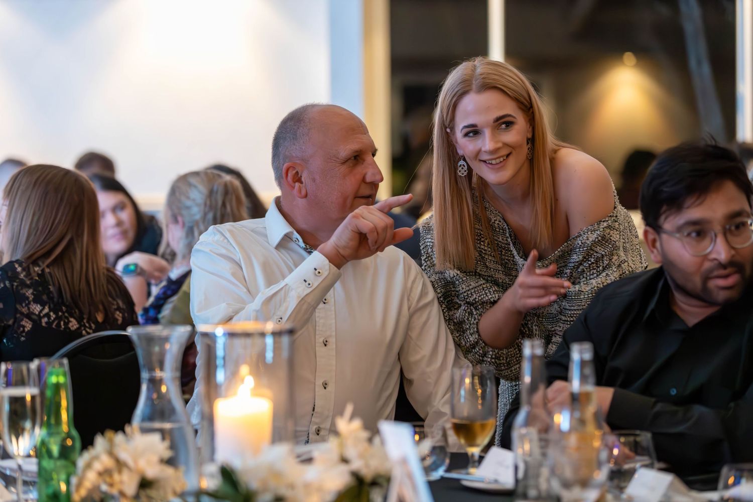 Guests socializing at an elegant dinner event, seated at a candlelit table with floral arrangements.