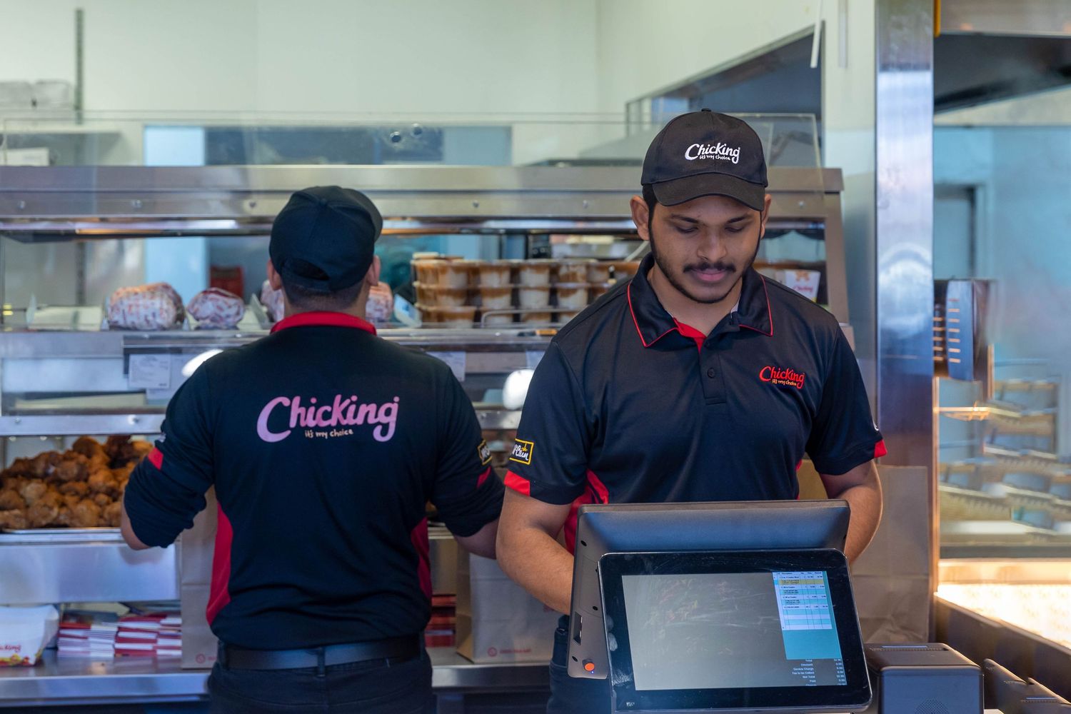 Two Chicking restaurant employees in branded uniforms working at the counter with a POS system and food display behind them.