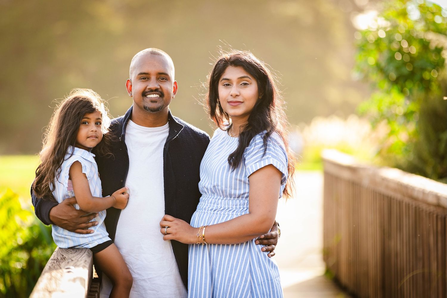 A happy South Asian family of three poses outdoors, with a smiling man holding a young girl beside a pregnant woman.