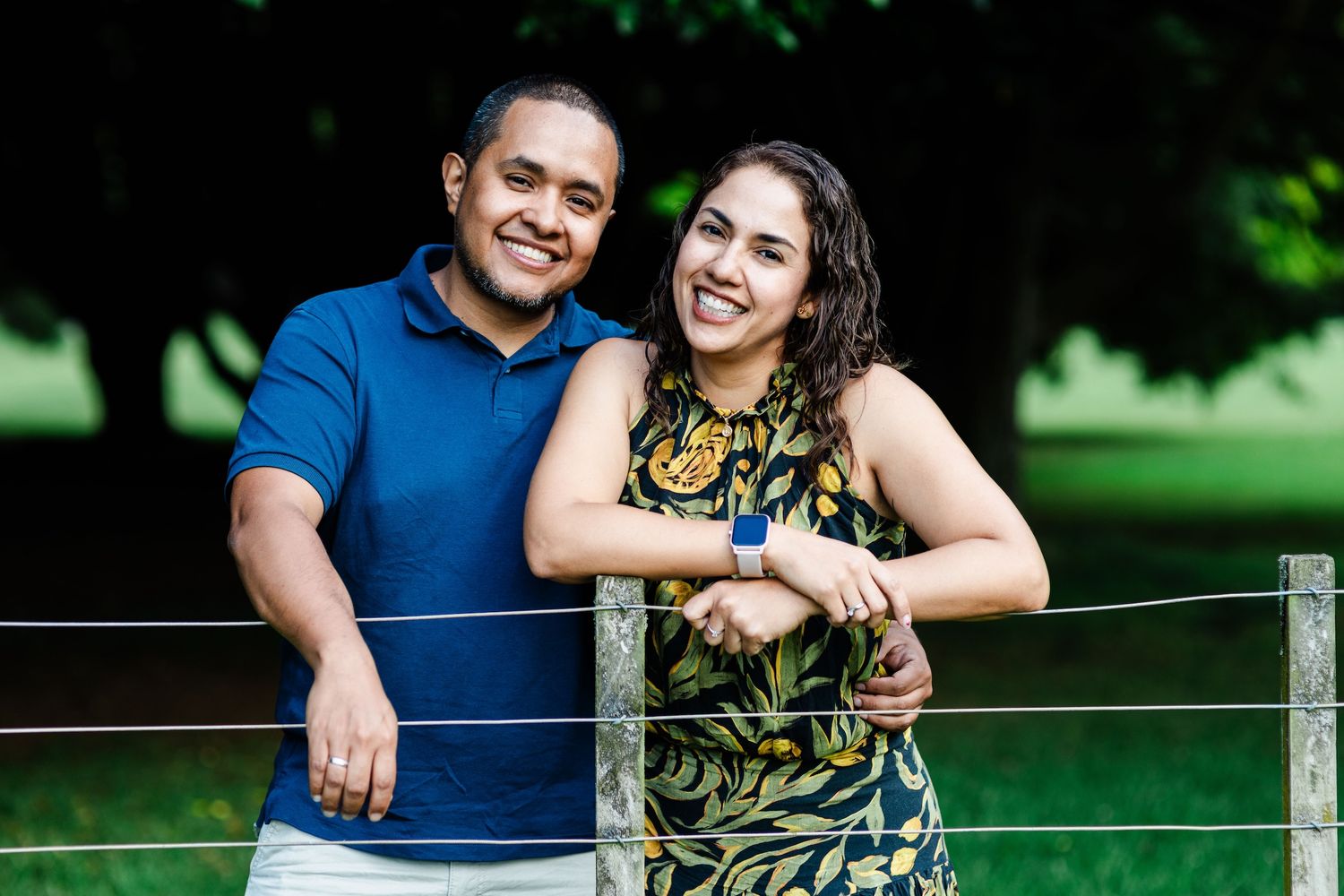 A smiling couple poses outdoors near a fence with lush green trees in the background on a sunny day.