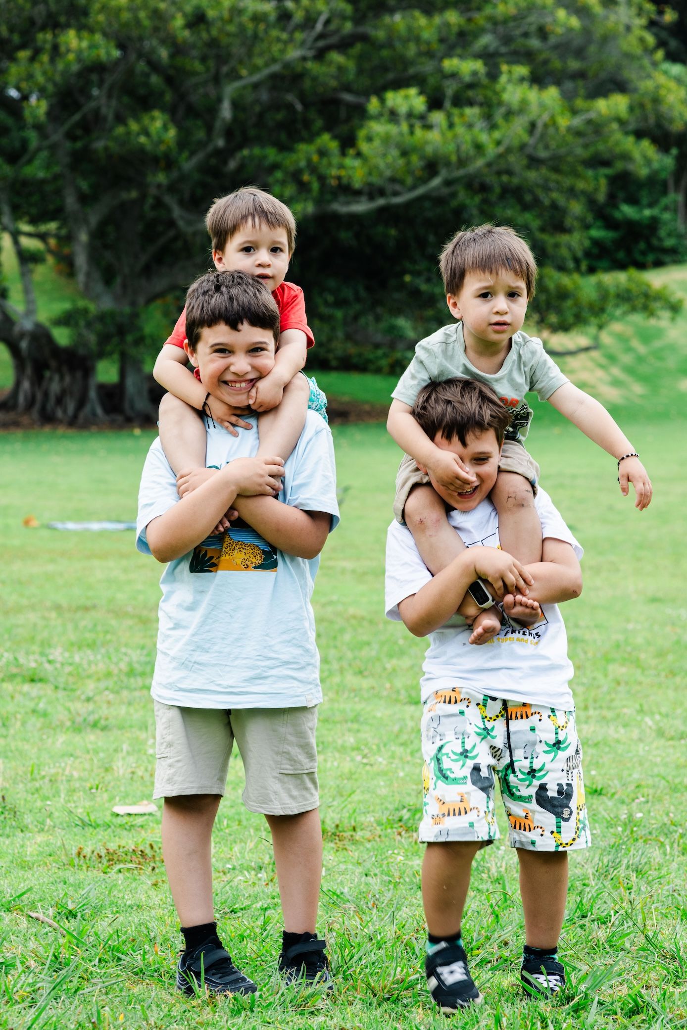 Four young boys playing outdoors, two older boys giving piggyback rides to younger toddlers on green grass.