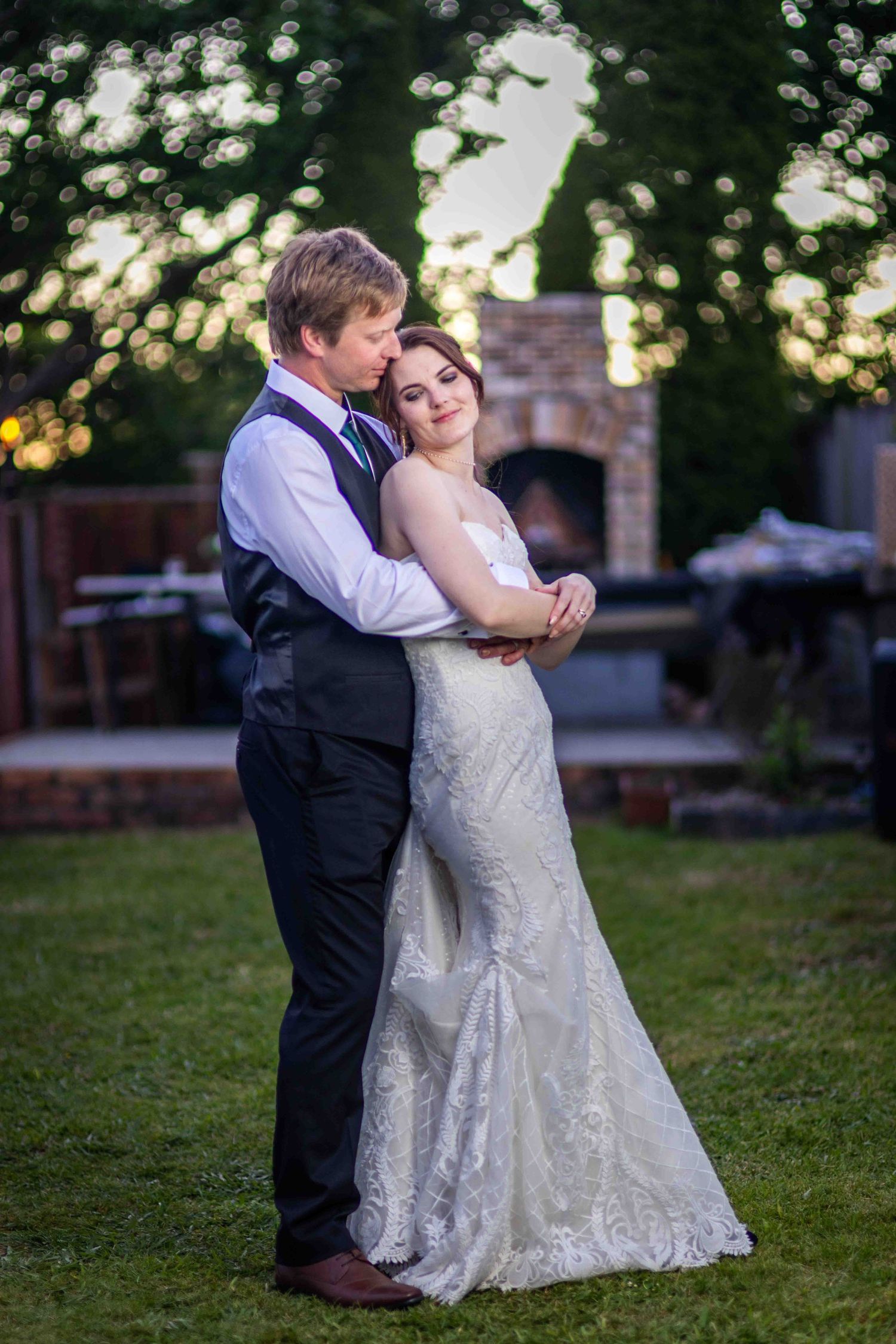 Bride and groom embracing outdoors at dusk, she in lace gown, he in dark vest, with brick fireplace backdrop.