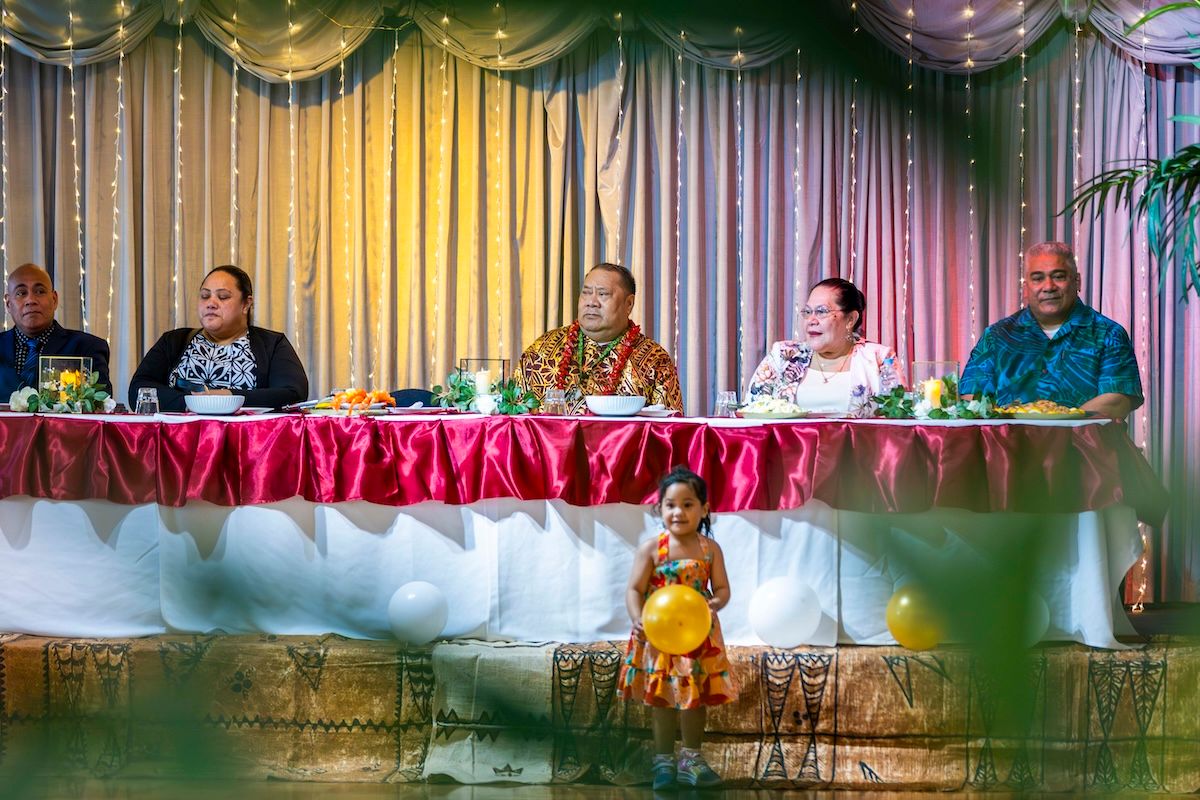 People seated at a long table with red tablecloth under decorative ceiling drapes and lights at a celebration.