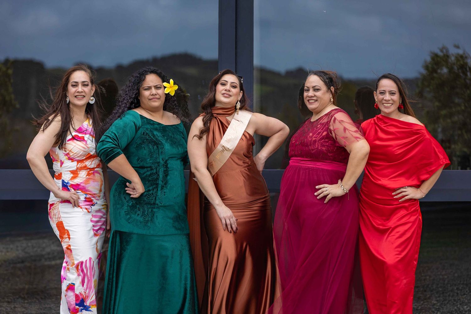 Five women in elegant formal gowns pose together outdoors, wearing vibrant colors including green, rust, red, and floral prints.