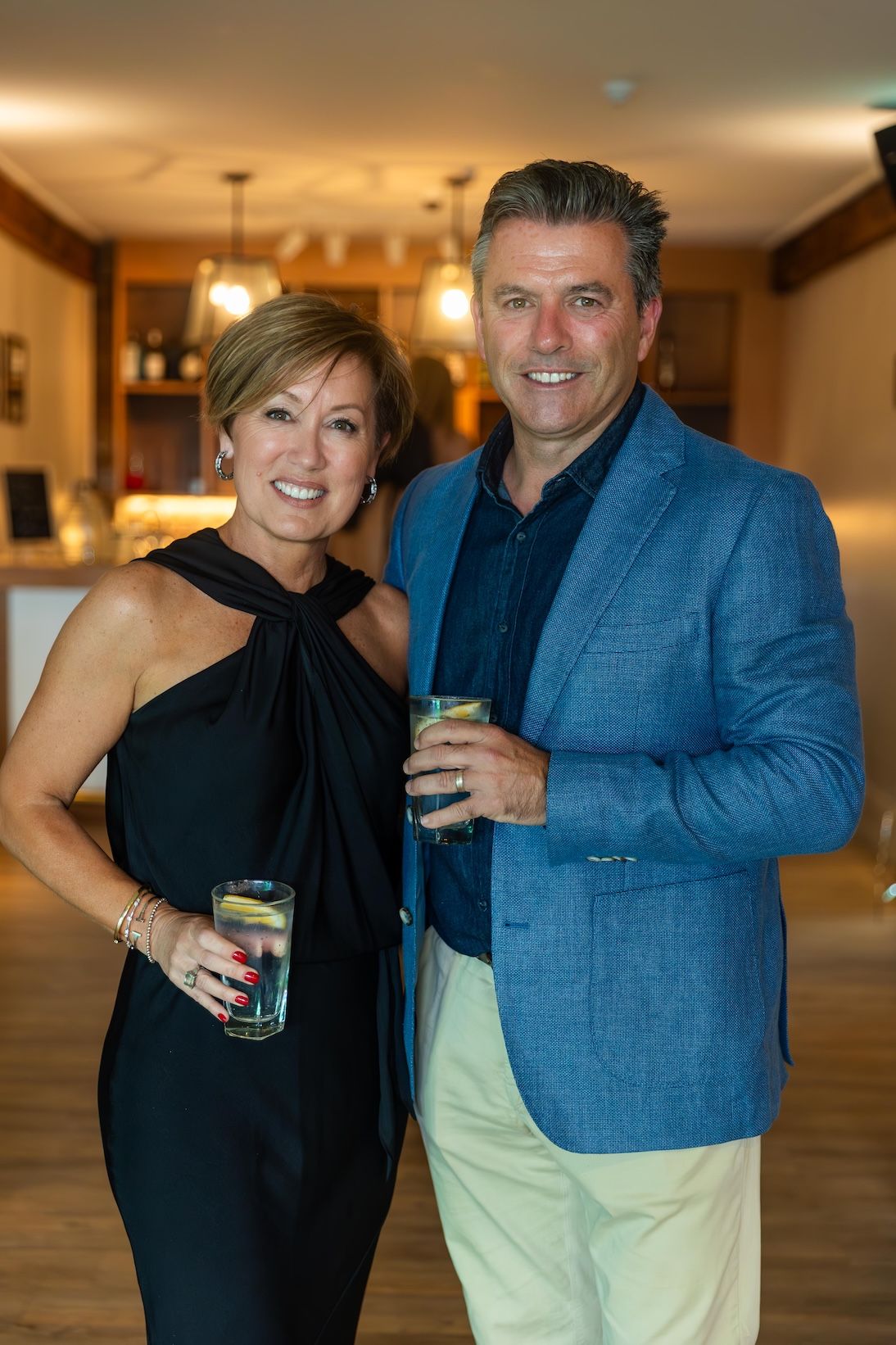 Smiling couple in elegant attire holding cocktails at an indoor social event.