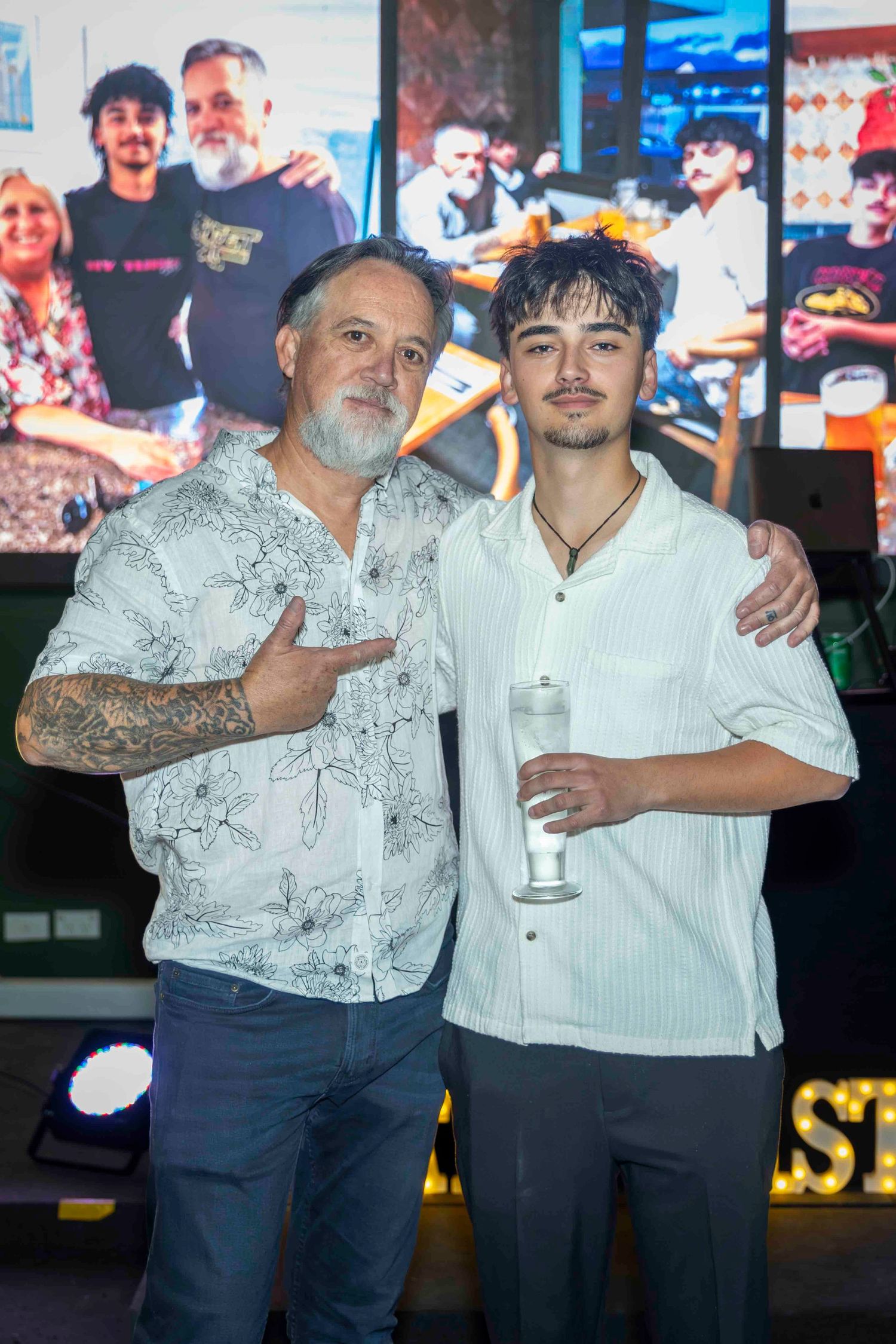 Two men posing together at an event, one in a floral shirt with tattoos, the other holding a drink.