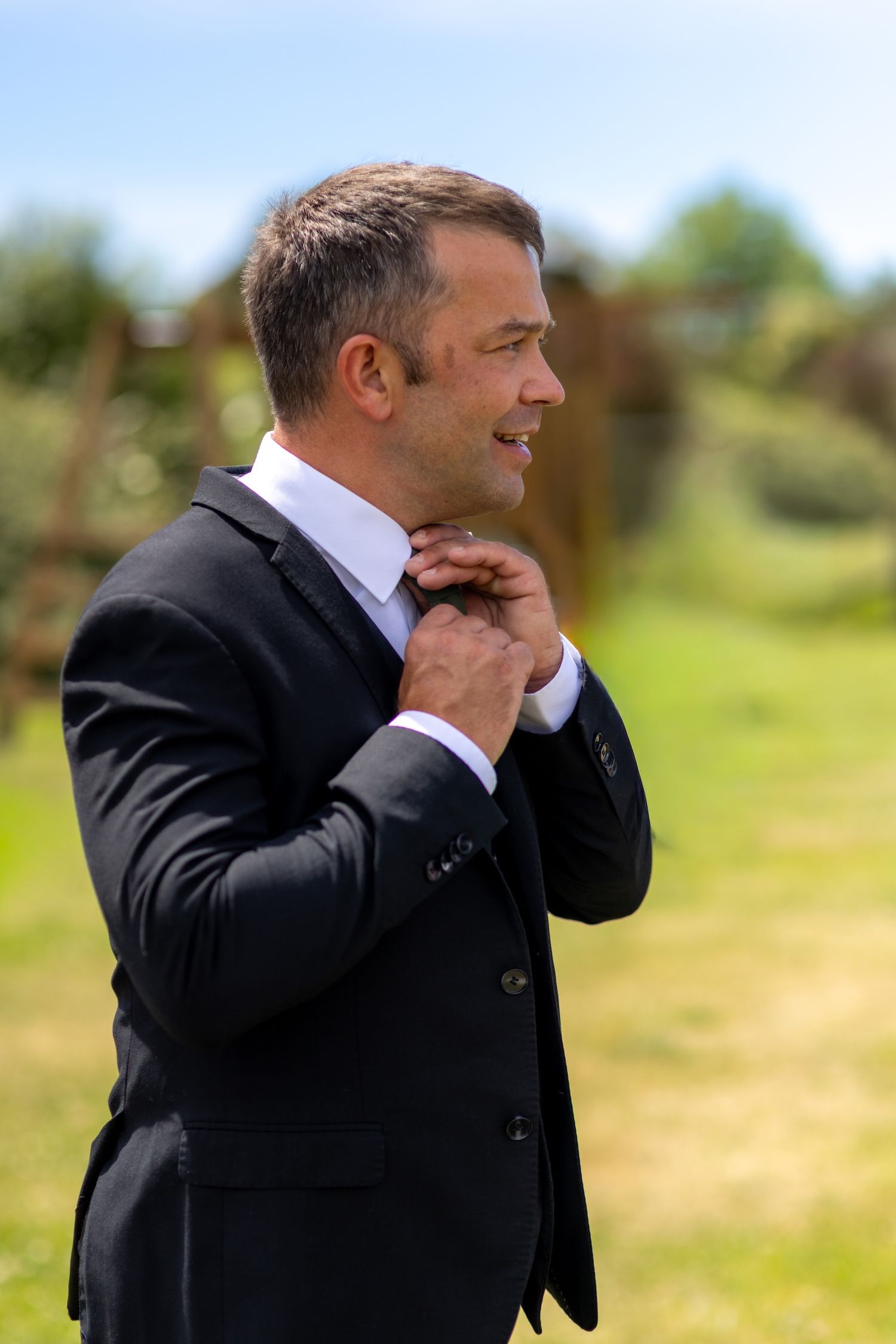 Elegant man in black suit adjusting collar outdoors on sunny day with green garden background.