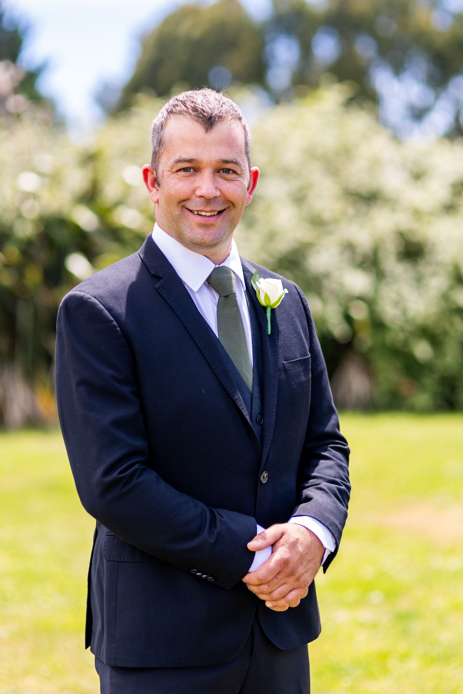 Smiling groom in navy suit with green tie and white boutonniere standing outdoors on a sunny day.