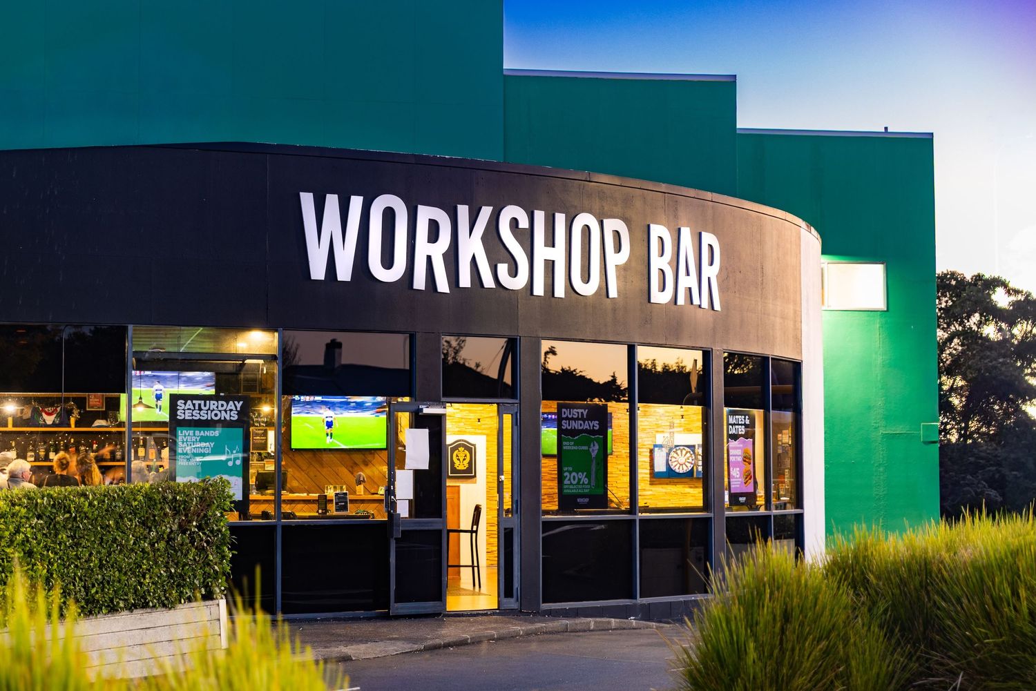 Modern Workshop Bar storefront illuminated at dusk with bright signage and green architectural accents.