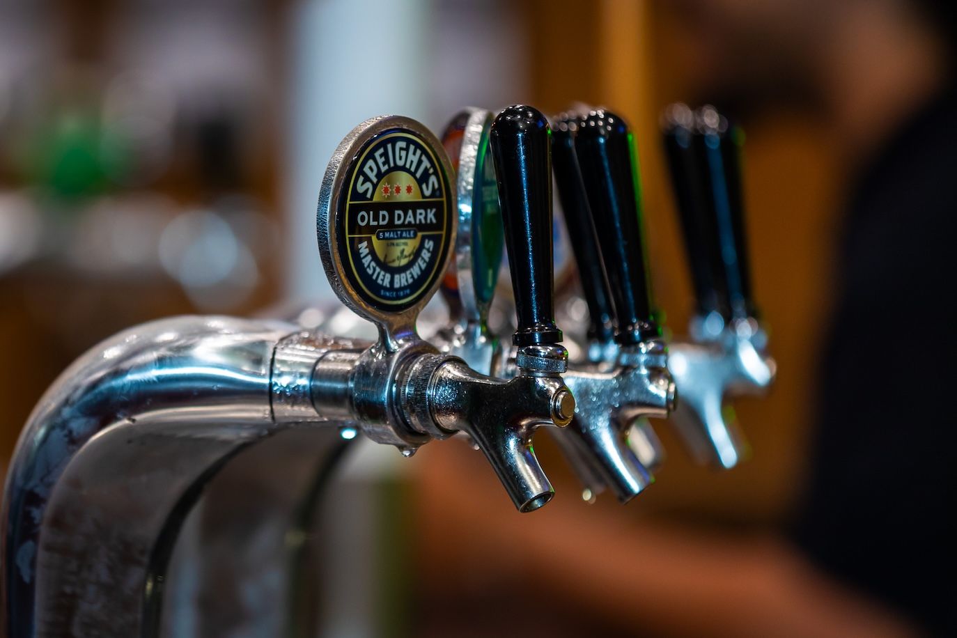 Row of chrome beer taps at a bar with blurred background showing bottles and glassware.
