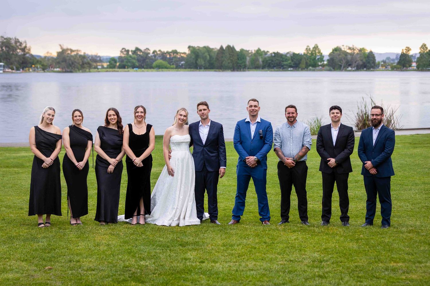 Wedding party lined up for formal group photo on grass with lake in background.