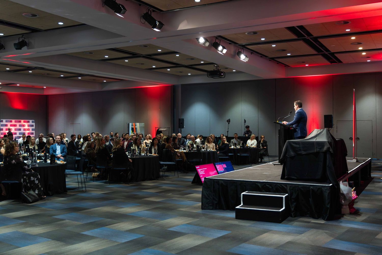 Conference hall with theater-style seating and dramatic red lighting during a presentation.