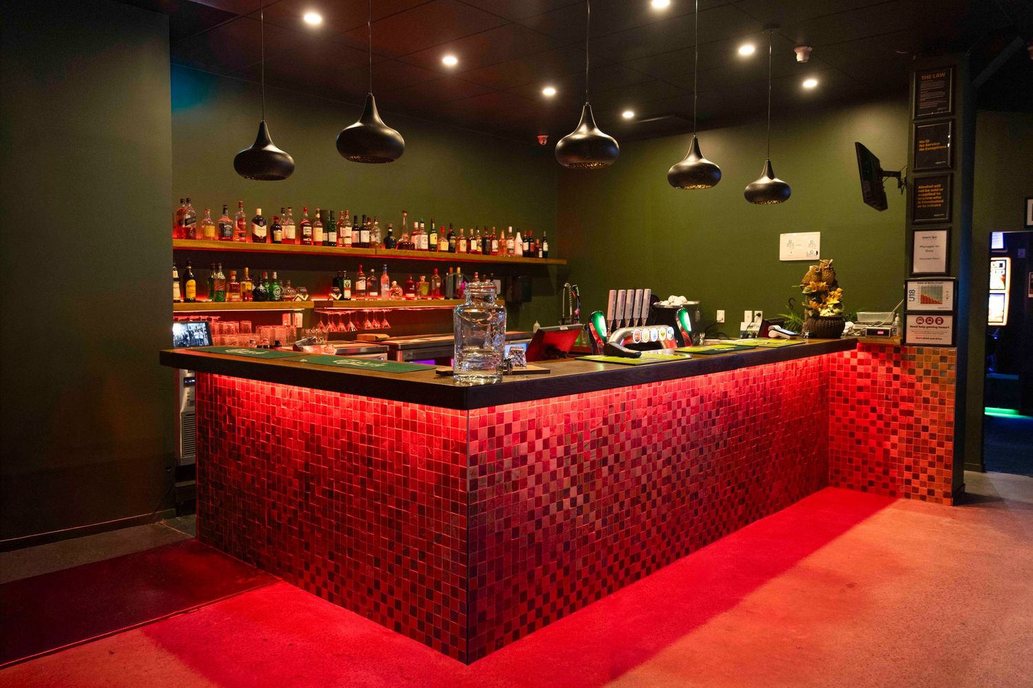 A stylish bar with a glowing red mosaic counter, dark green walls, and pendant lights illuminating shelves of spirits.