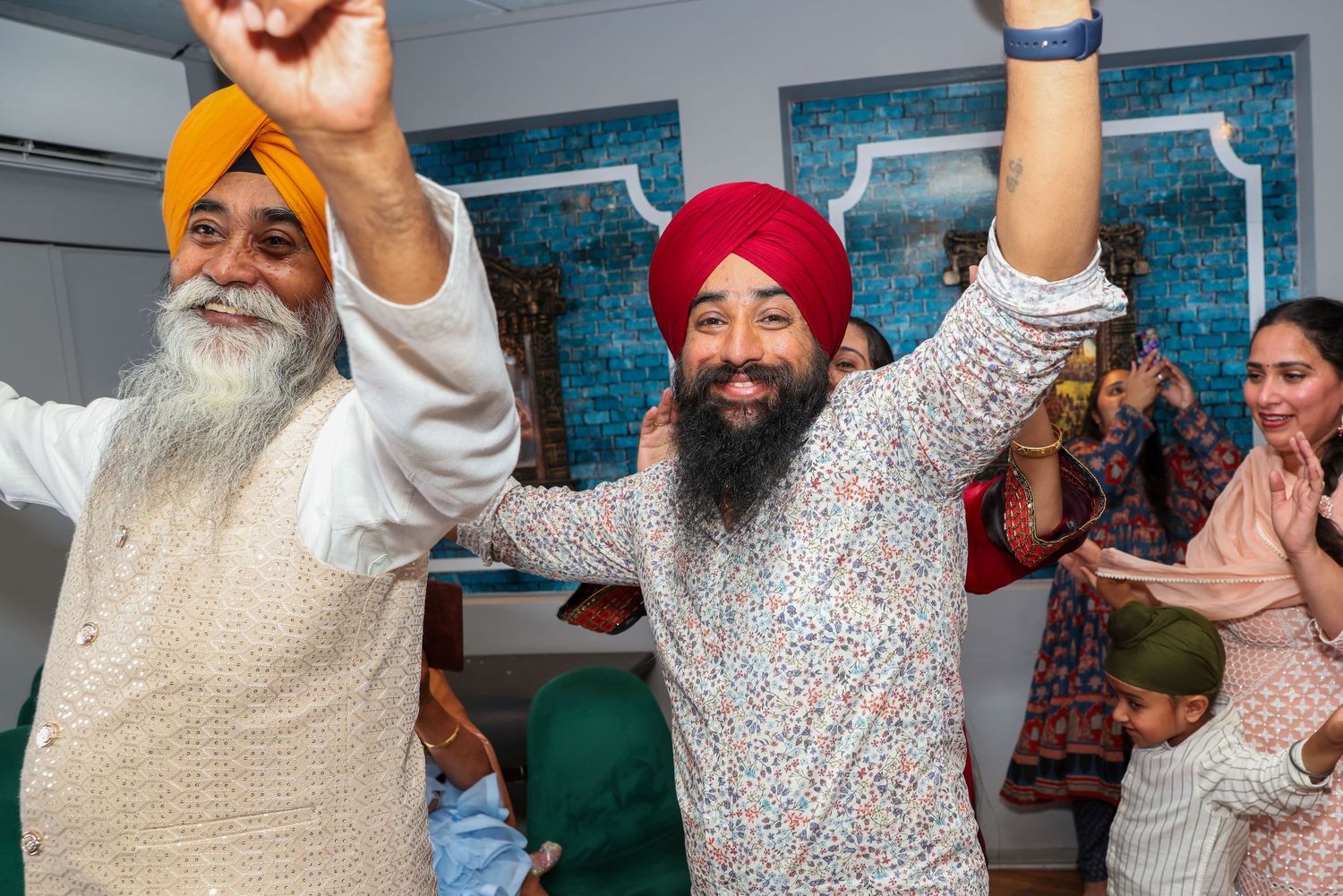 Two Sikh men in colorful turbans celebrate joyfully with raised hands at a festive gathering.