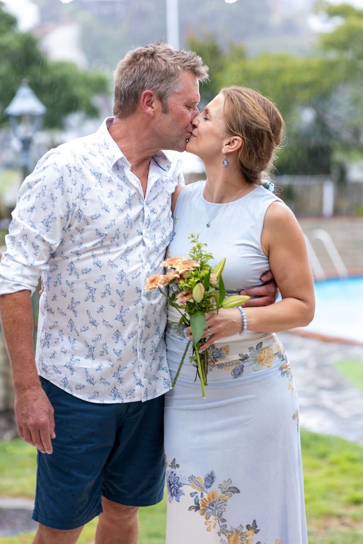 Middle-aged couple sharing a romantic kiss outdoors, woman holding bouquet of peach roses, near a pool.