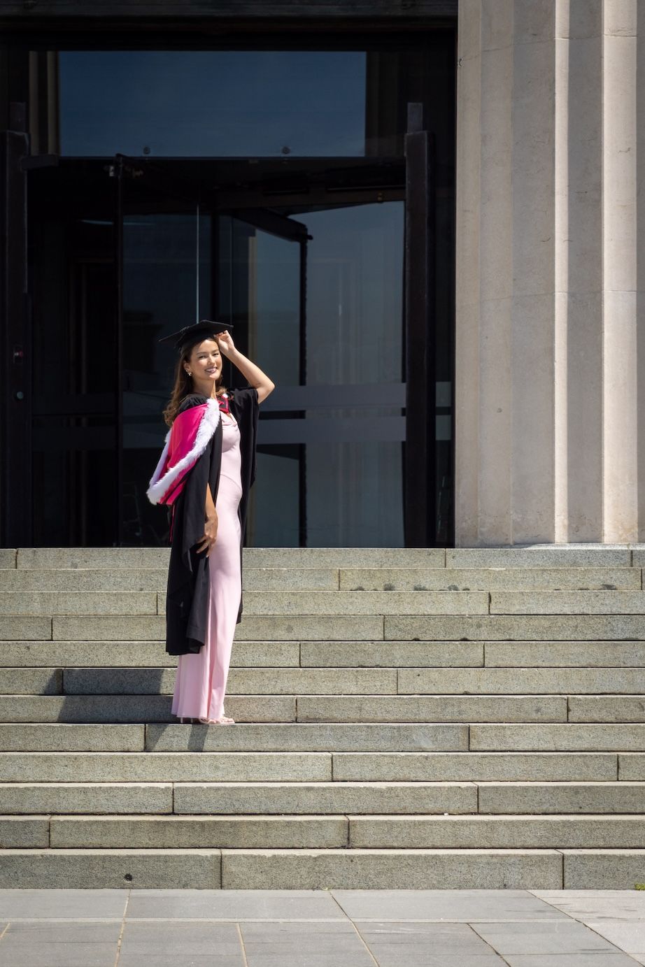 A smiling graduate in black cap and gown with pink sash poses on stone steps outside a university building.