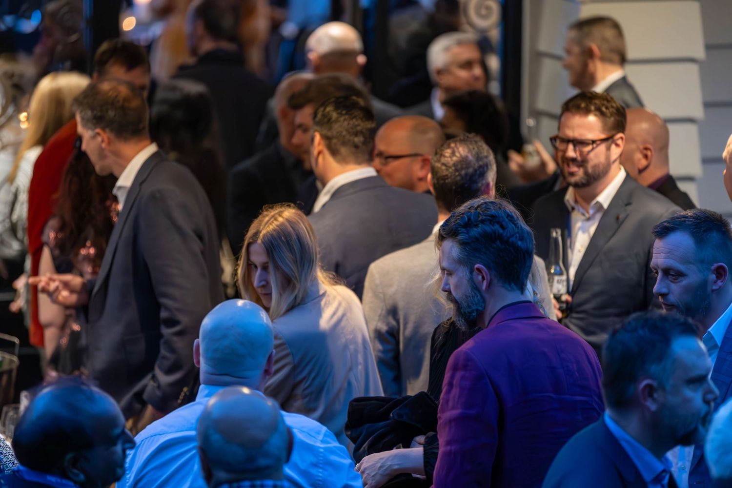 Professionals networking at a corporate event, mingling in a crowded venue with blue ambient lighting.