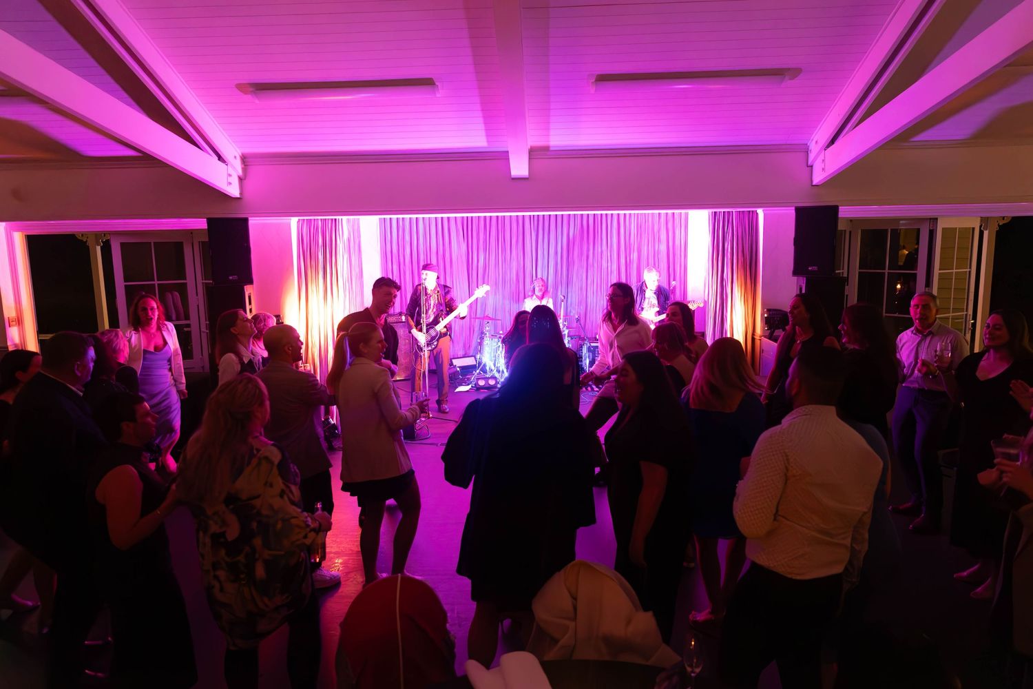 Guests dancing at an indoor event with pink and purple uplighting and a live band performing on stage.