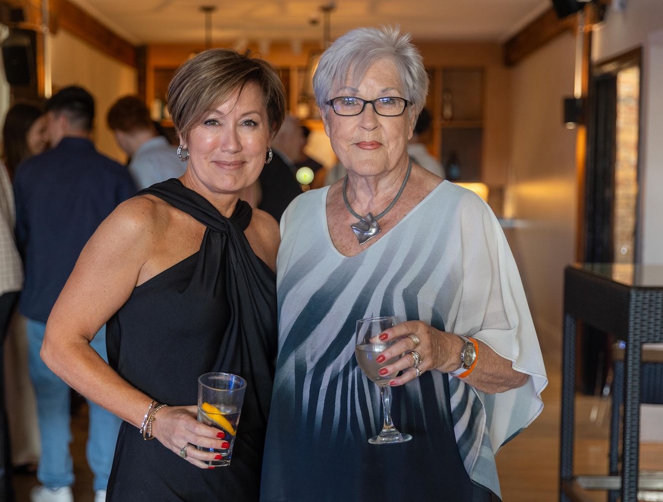 Two women in elegant attire smiling and holding cocktails at an indoor social event.