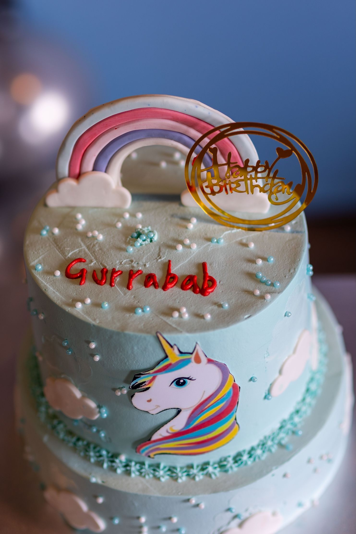 A two-tier light blue unicorn birthday cake with rainbow topper, clouds, and Happy Birthday sign for Gurrabab.