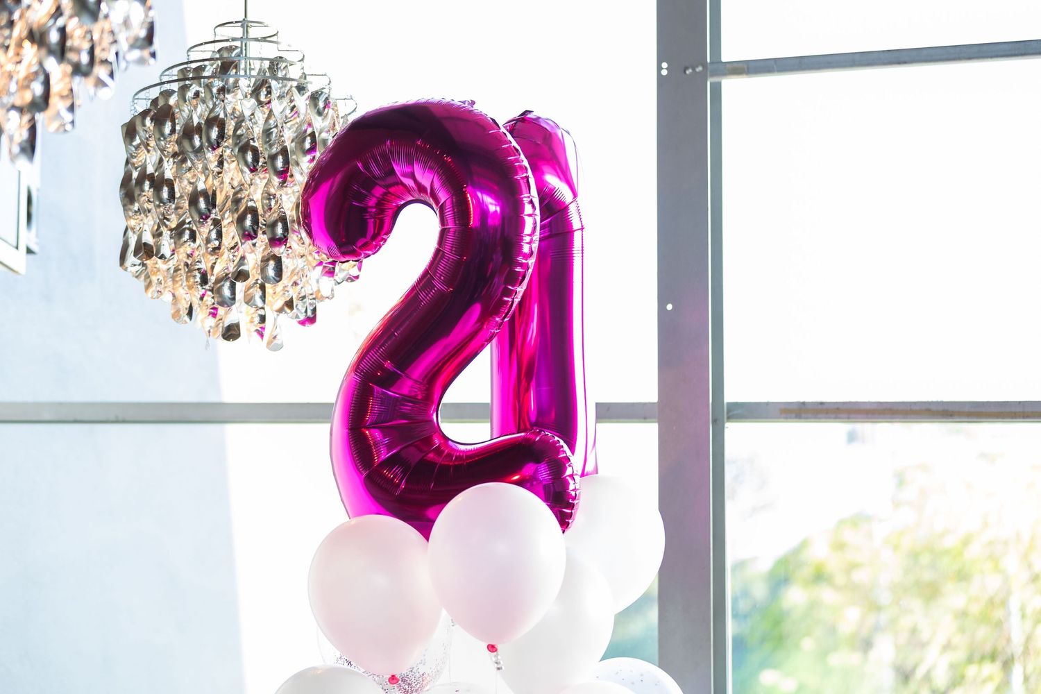Pink foil number 21 birthday balloons with white balloons on floor near window and chandelier.