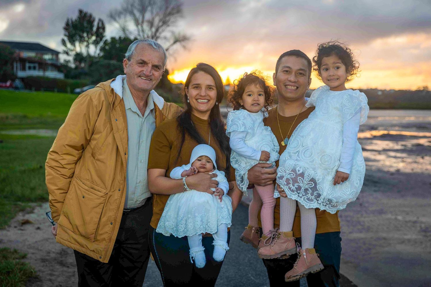 Family of five posing outdoors at sunset near a lake, with two adults holding babies in white dresses.
