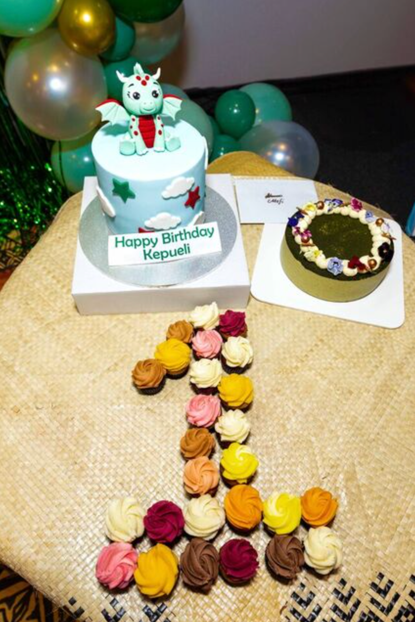 First birthday celebration with dragon-themed cake reading Happy Birthday Kepueli, colorful cupcakes arranged in number one shape.