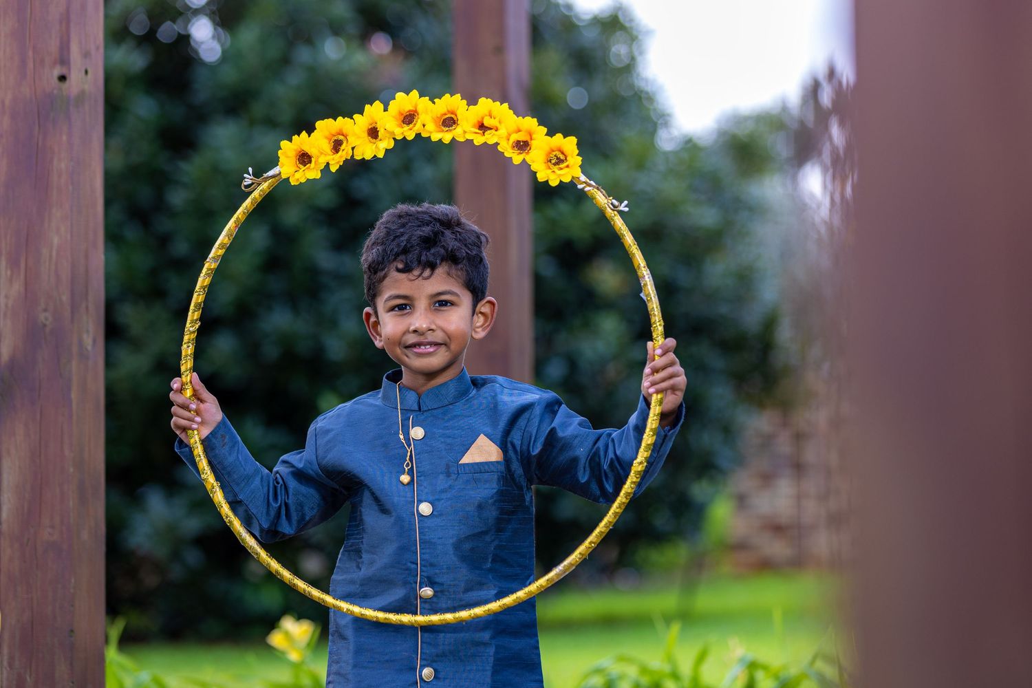 Young boy in blue kurta smiling through a yellow flower-decorated hula hoop in a garden setting.