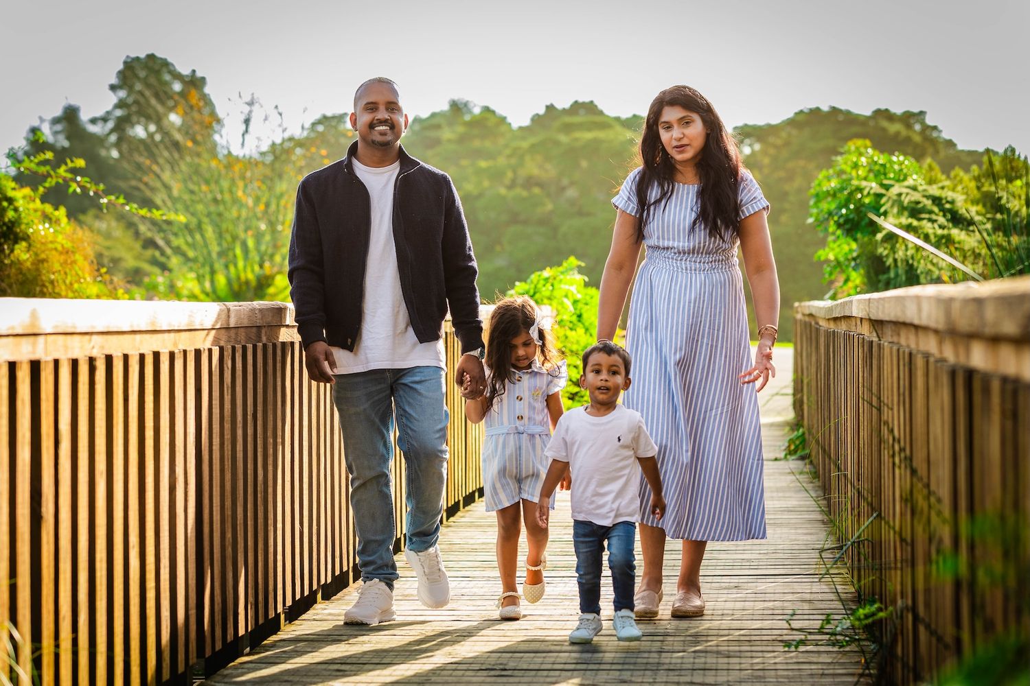 Family of four walking on a wooden bridge path surrounded by lush greenery in warm golden sunlight.