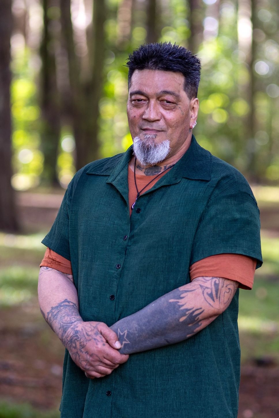 Tattooed man with gray beard in green shirt stands outdoors among trees, smiling confidently.