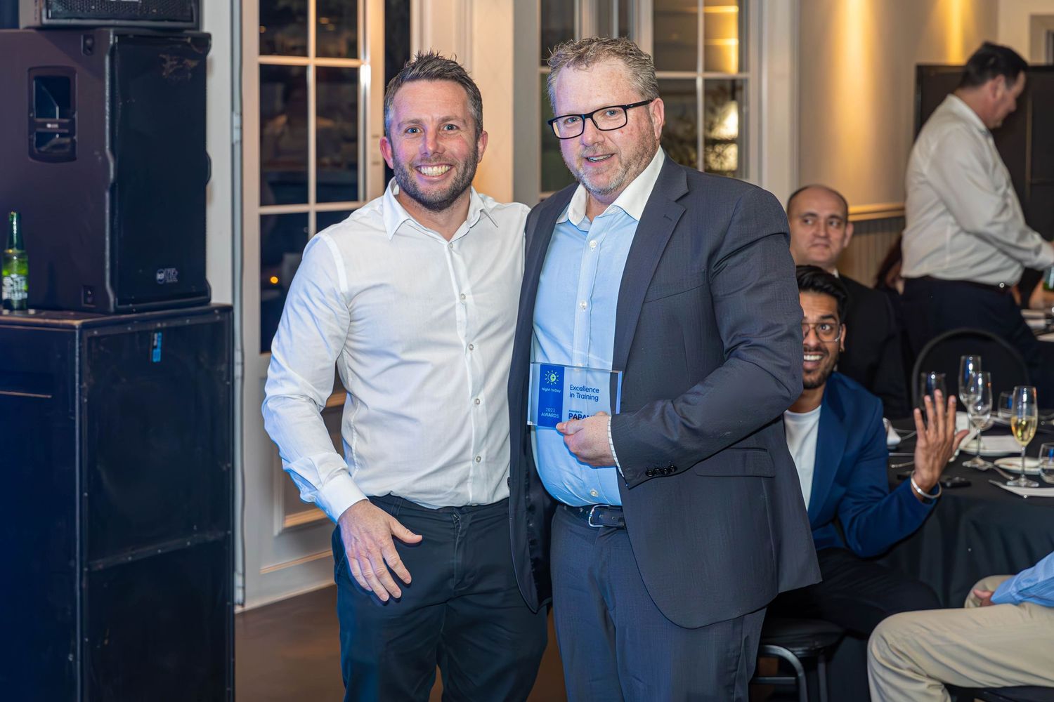 Two men pose together at an indoor corporate awards event, one holding a blue award plaque.