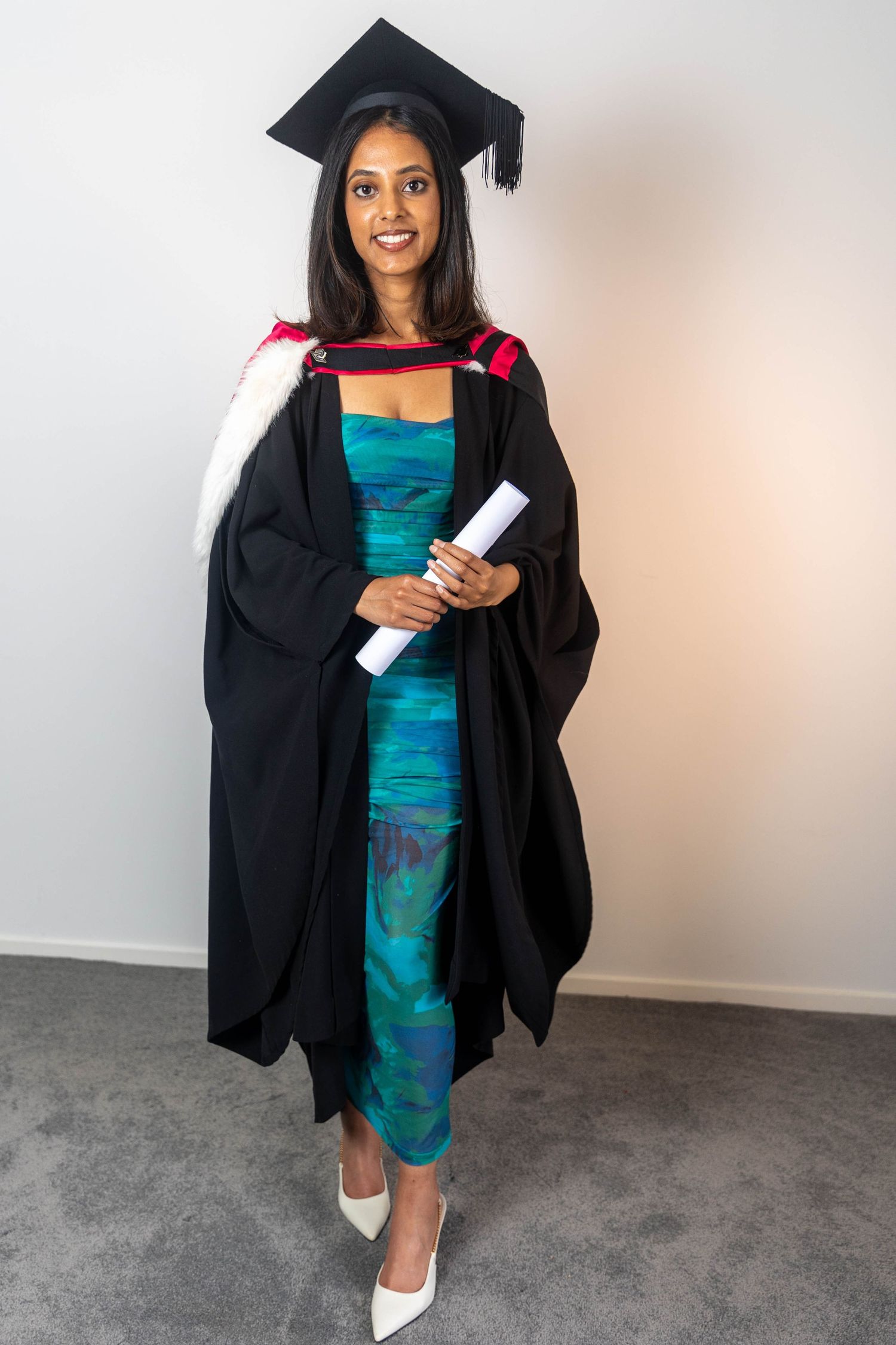 Smiling woman in black graduation gown and cap holding a diploma scroll, wearing a teal dress and white heels.