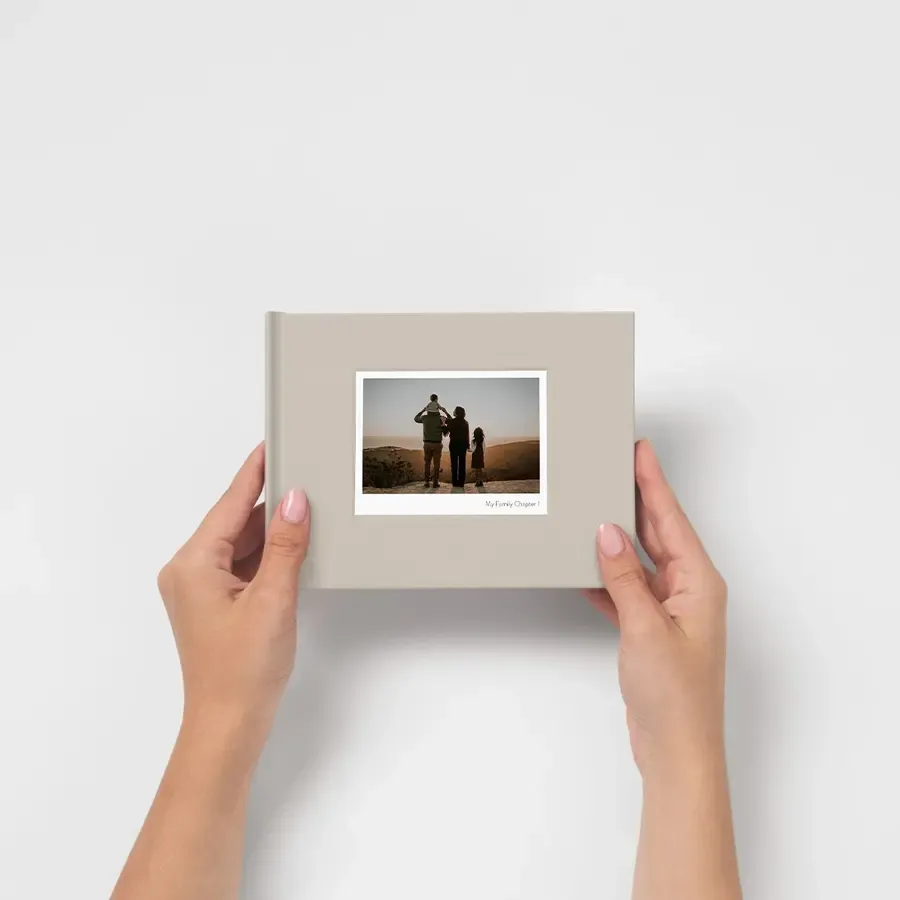 Hands holding a small hardcover photo book with a beige linen cover featuring a printed family portrait photo.