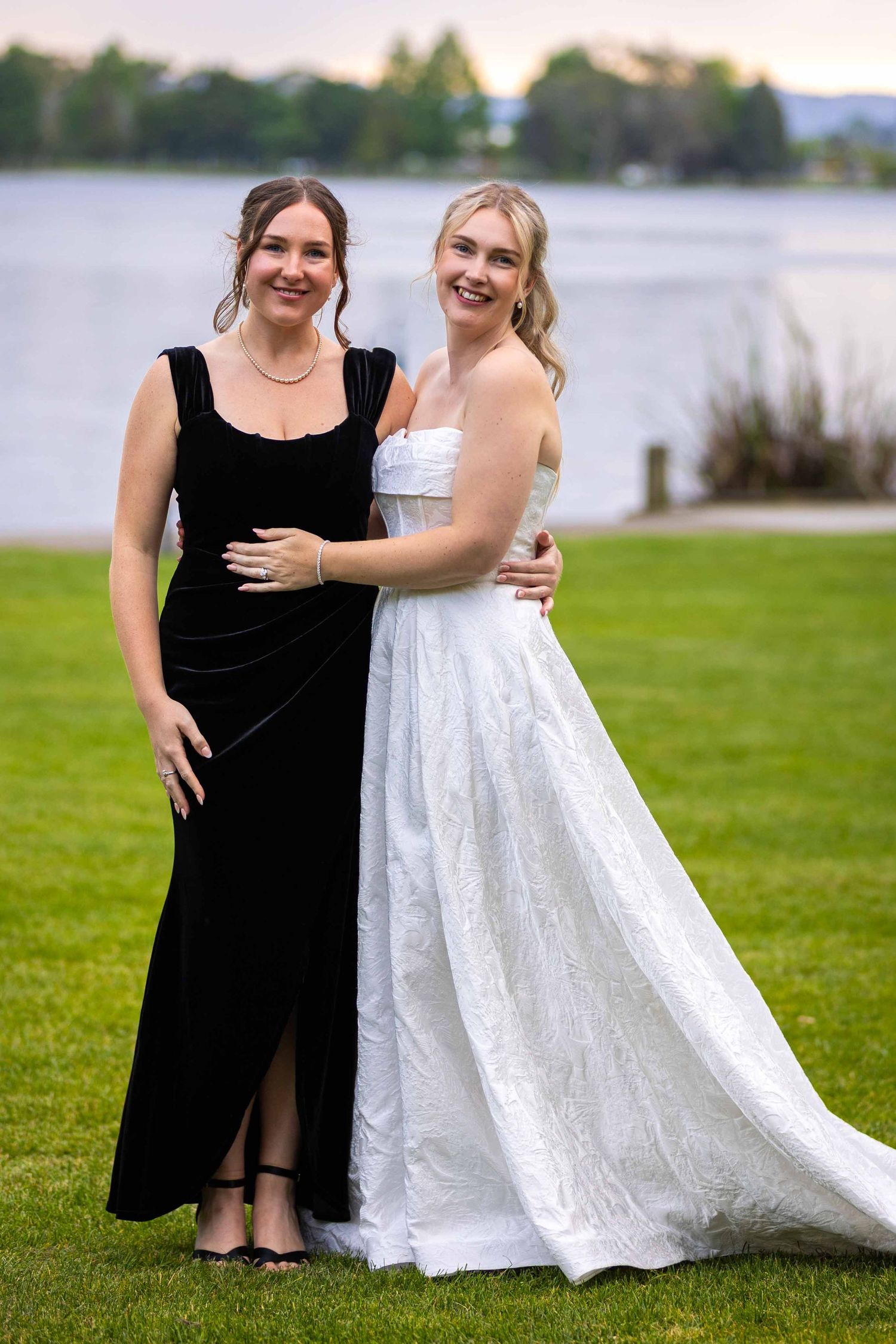 Two people in formal attire pose together on grass with a lake in the background.