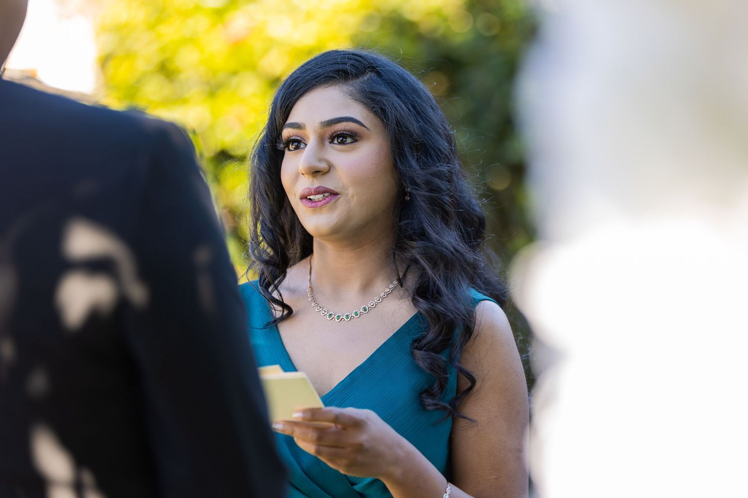 Young woman in teal dress holding a card, wearing a silver necklace, at an outdoor formal event.