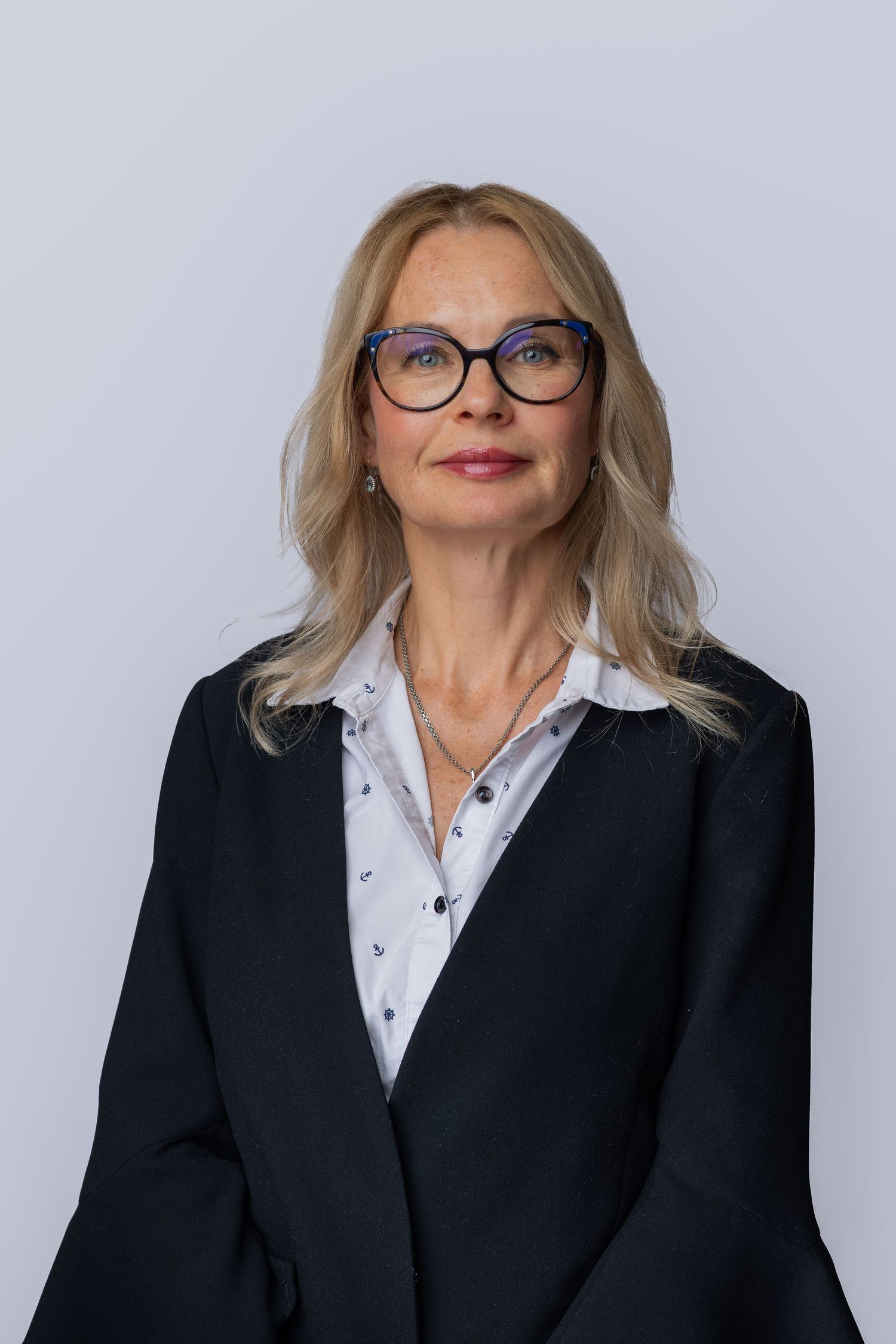 Professional businesswoman with blonde hair and glasses wearing a dark blazer over a white shirt poses for a portrait.