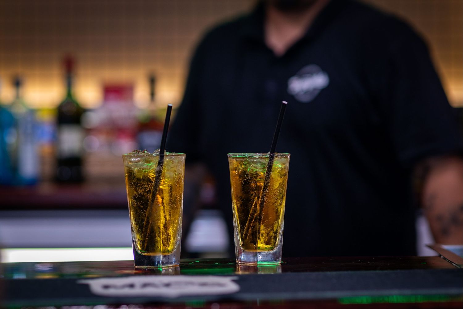 Two tall glasses of golden cocktails with ice sit on a dark bar counter while a bartender stands in the background.