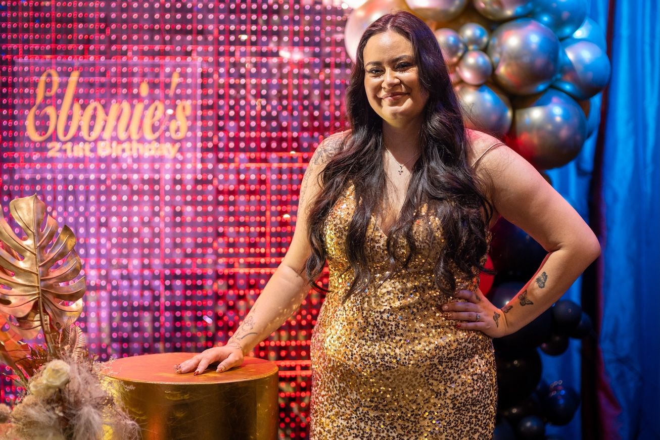 Woman in gold sequin dress posing at Ebonie's glamorous event with balloon backdrop and tropical decor.