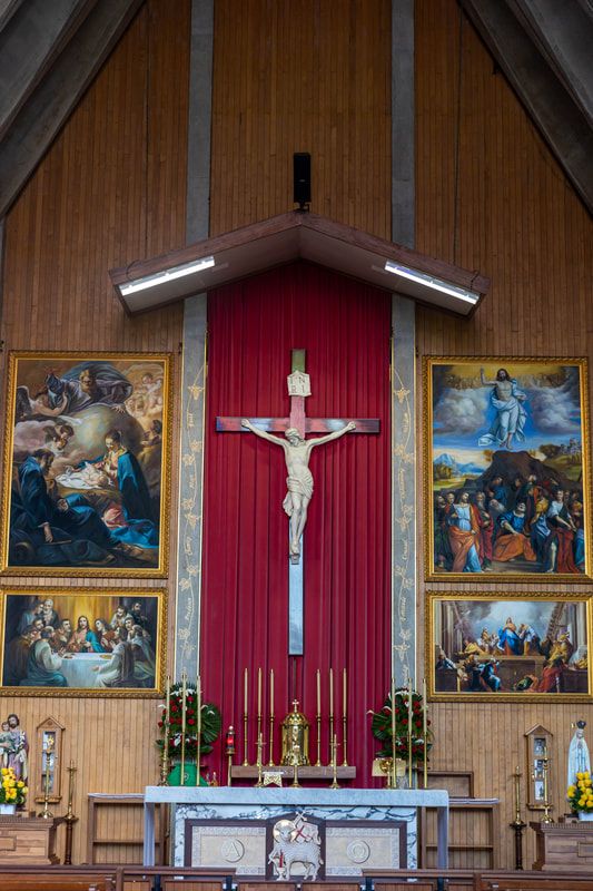 Catholic church altar with crucifix, red backdrop, religious paintings, candles, and floral arrangements in a wooden interior.